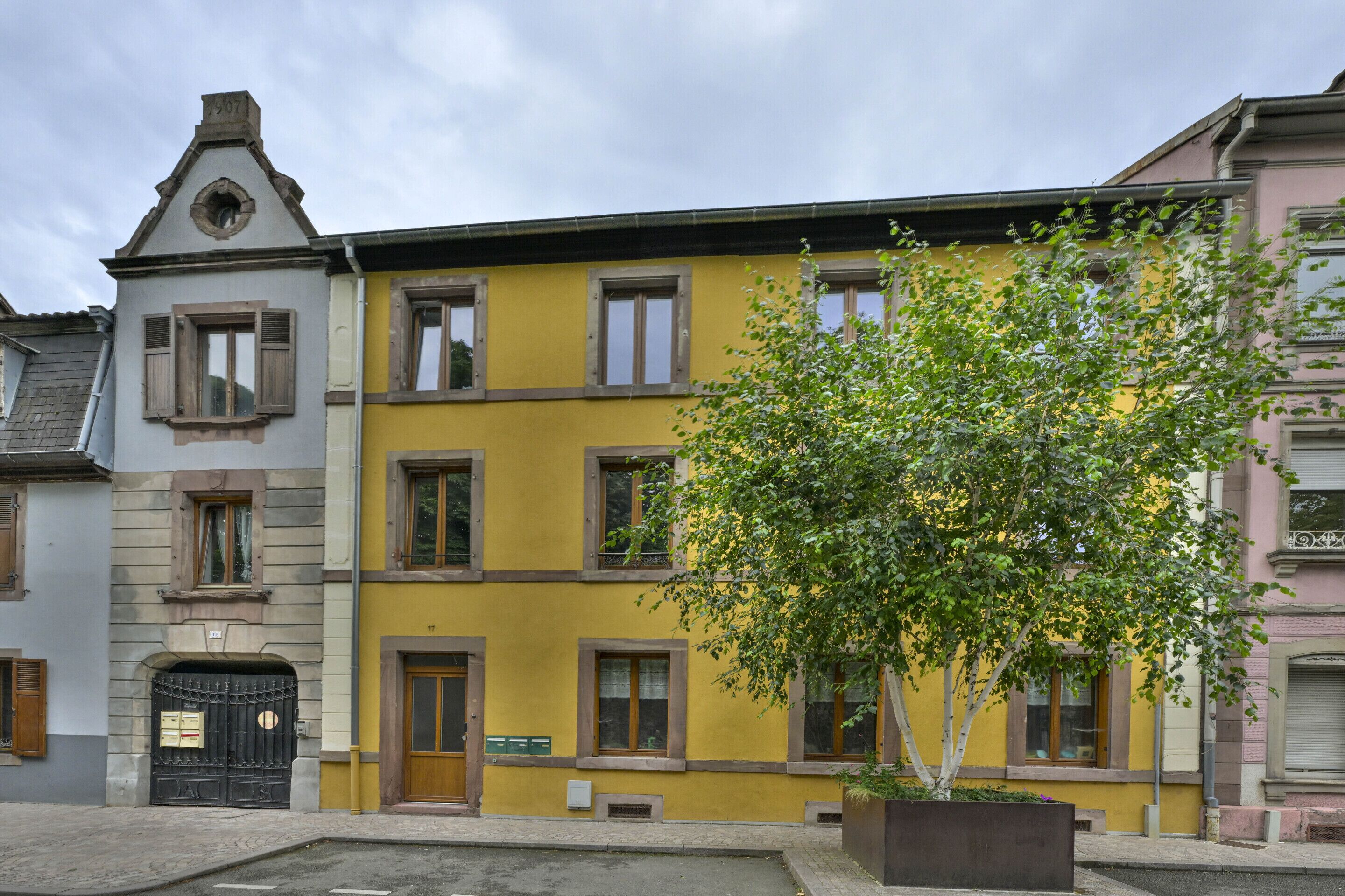 A multi-story building with a yellow facade, a tree in the foreground, and a black gate with mailboxes.