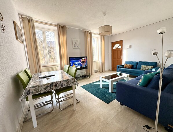 Una sala de estar con televisor, un sofá azul, una mesa de comedor con sillas y una mesa auxiliar.