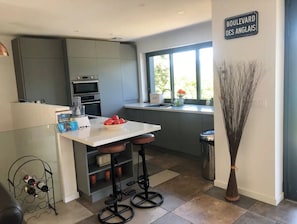 Une cuisine moderne avec un plan de travail blanc, deux tabourets de bar, un meuble à bouteilles et une fenêtre donnant sur un paysage verdoyant.