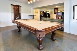 Uma mesa de bilhar em um espaço com um sofá e jogos de fliperama ao fundo.