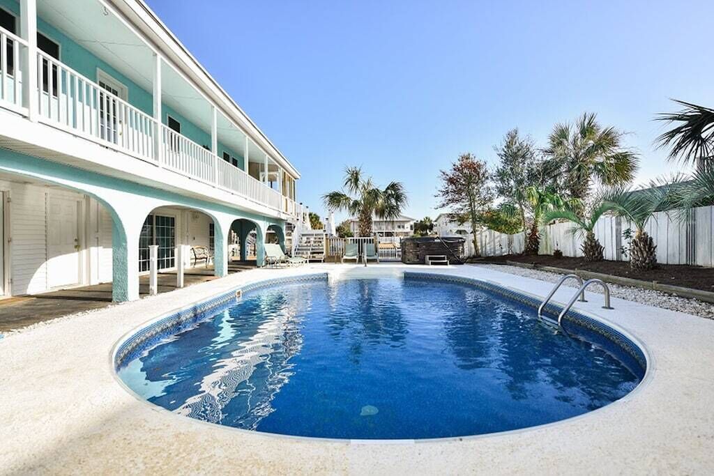 A swimming pool with a white deck, a covered patio with white railings, and palm trees in the background.