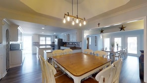 Uma sala de jantar espaçosa com uma mesa de madeira e cadeiras brancas, cozinha ao fundo e um lustre.