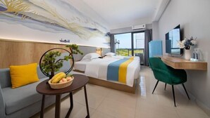 Habitación de hotel con cama, escritorio, silla, mesita con fruta, planta y ventana grande.
