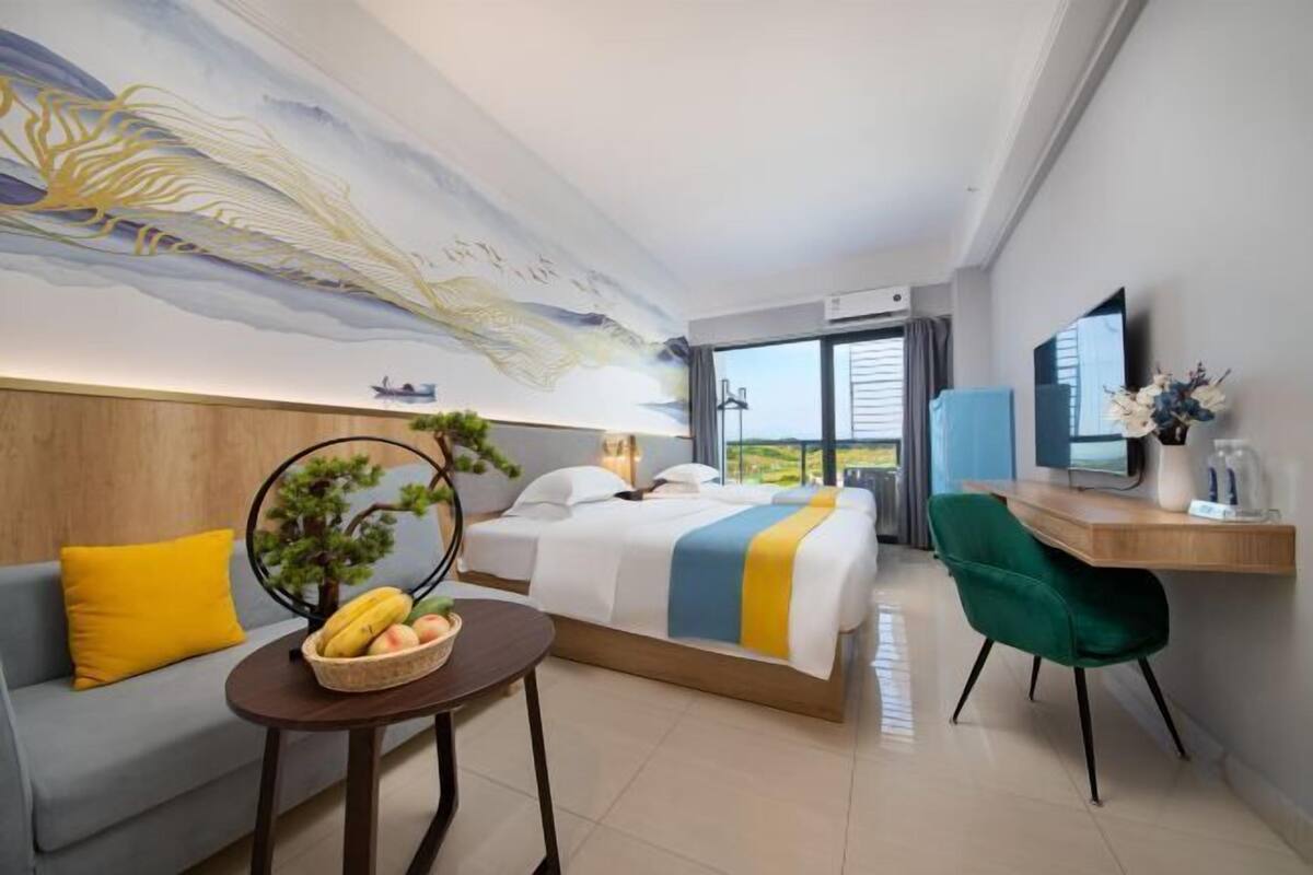 Habitació d'hotel amb un llit, una taula de treball, una cadira, una taula petita amb fruita, una planta i una finestra gran.