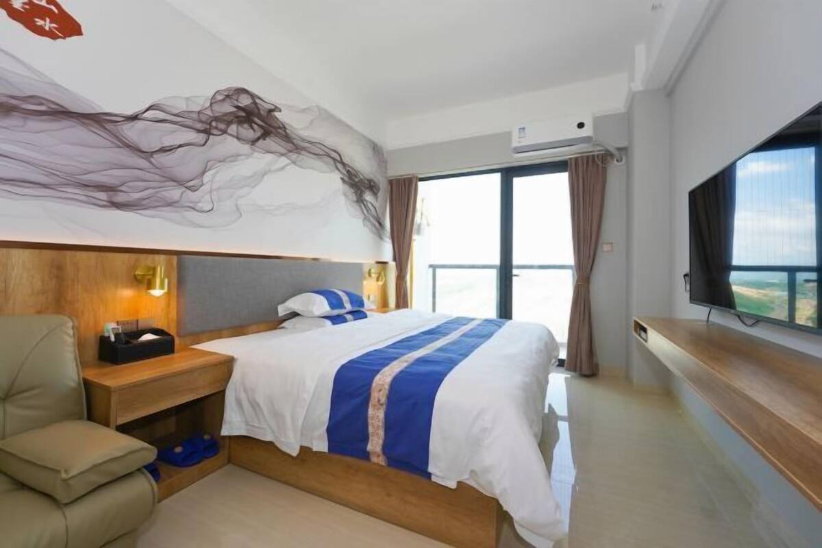Habitació d'hotel amb llit, una nit, una cadira, un televisor i una finestra gran.
