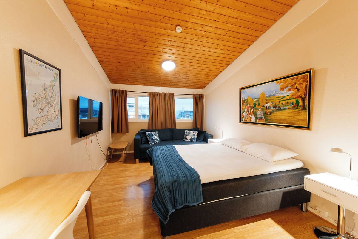 A hotel room with a wooden ceiling, a bed, a desk, a TV, a painting, and a map on the wall.