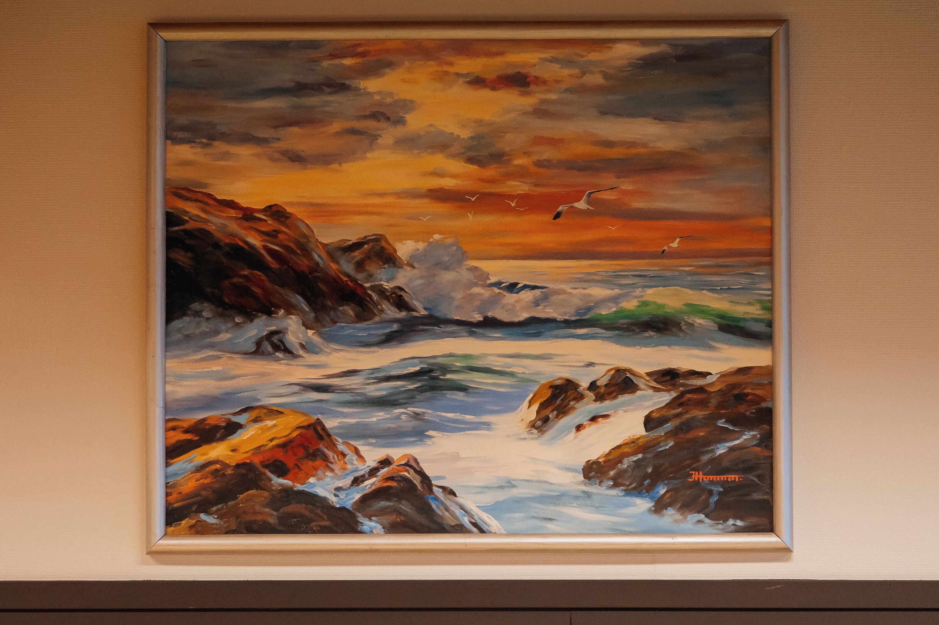 A framed painting of a seascape with waves and rocks under a sunset sky.