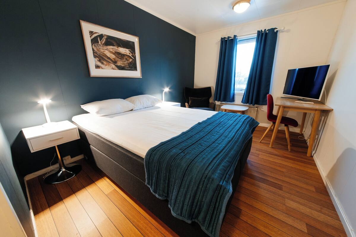 A hotel room with a bed, bedside tables, a desk with a chair, a television, and a window with curtains.