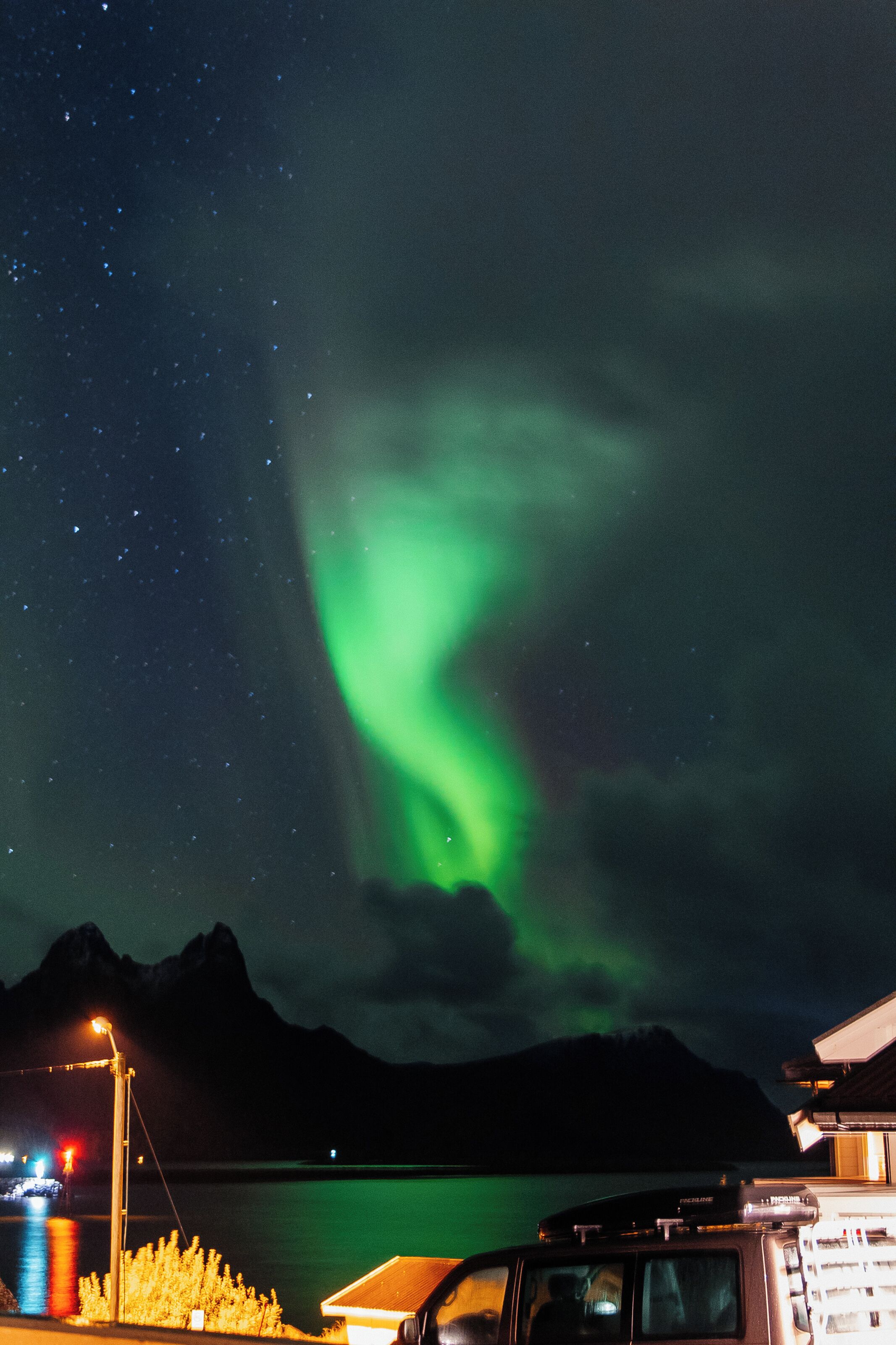 A night scene with the Northern Lights over a body of water, mountains in the background, and a building with a car parked in front.