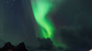 A night scene with the Northern Lights over a body of water, mountains in the background, and a building with a car parked in front.