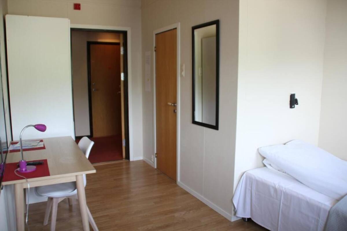 A room with a wooden floor, a desk with a chair, a bed, and a door.