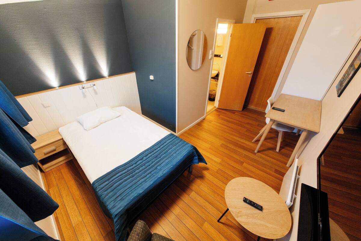 A compact hotel room with a single bed, a small table, a TV, and a door leading to a bathroom.