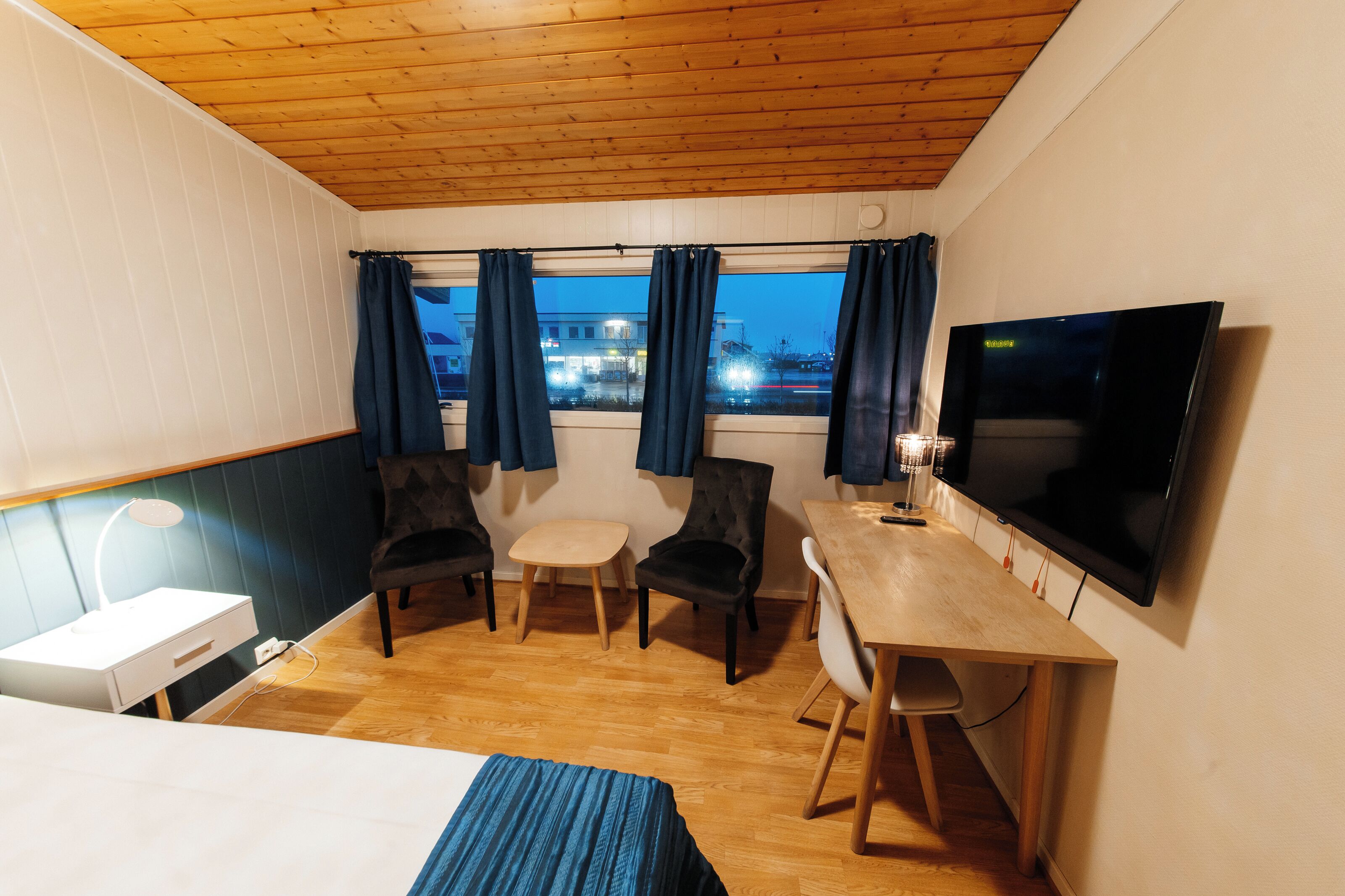 Une chambre d’hôtel compacte avec un plafond en bois, une grande télévision, un bureau avec des chaises, une petite table et un lit recouvert d’une couverture à rayures bleues.