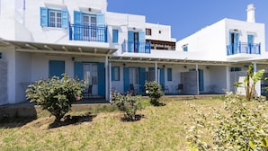 Un edificio blanco con contraventanas azules y balcones, un área de estar al aire libre techada y pequeños arbustos.