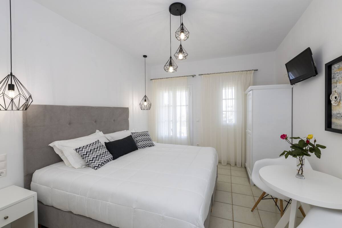 A modern bedroom with a large bed, bedside tables, a small round table with a vase, and a flat-screen TV mounted on the wall.