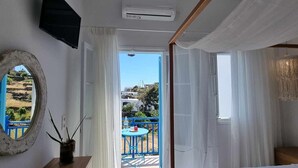 Un dormitorio con cama, escritorio con silla, televisor y aire acondicionado. Se ve un área exterior azul con mesa y sillas.
