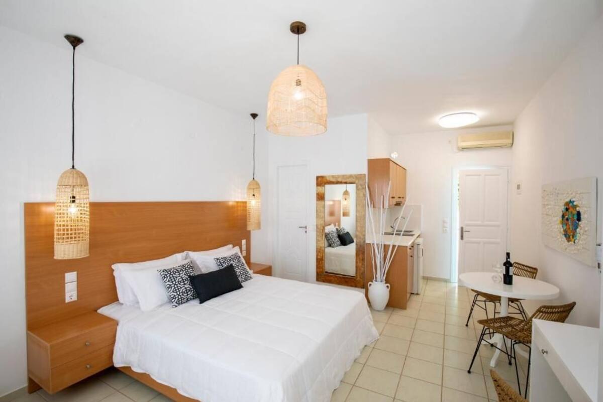 A bedroom with a bed, bedside tables, a mirror, a dining area with a table and chairs, and a kitchenette.