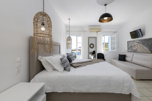 A modern bedroom with a large bed, a sofa, a TV, and a hanging light fixture. - Eutuxia Rooms & Studios (Sifnos)