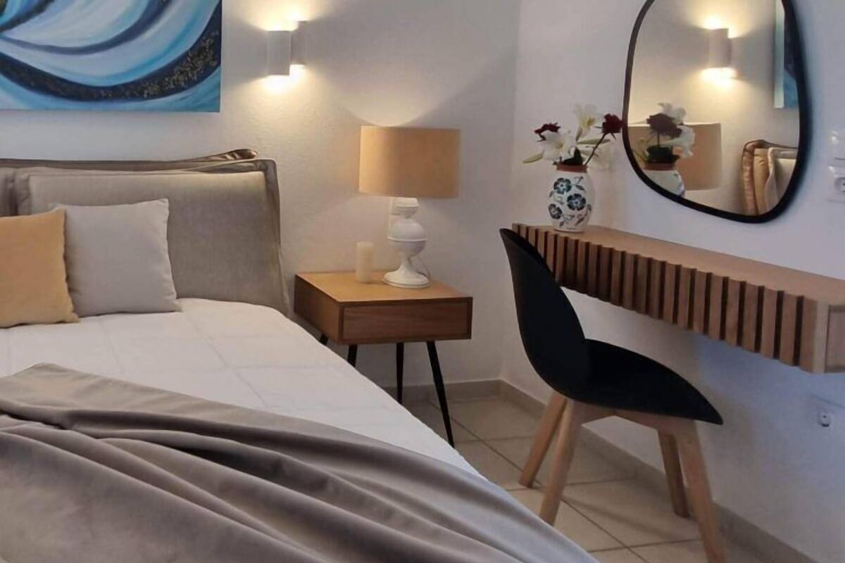 A hotel room with a bed, a desk, a chair, a lamp, a mirror, and a painting on the wall.