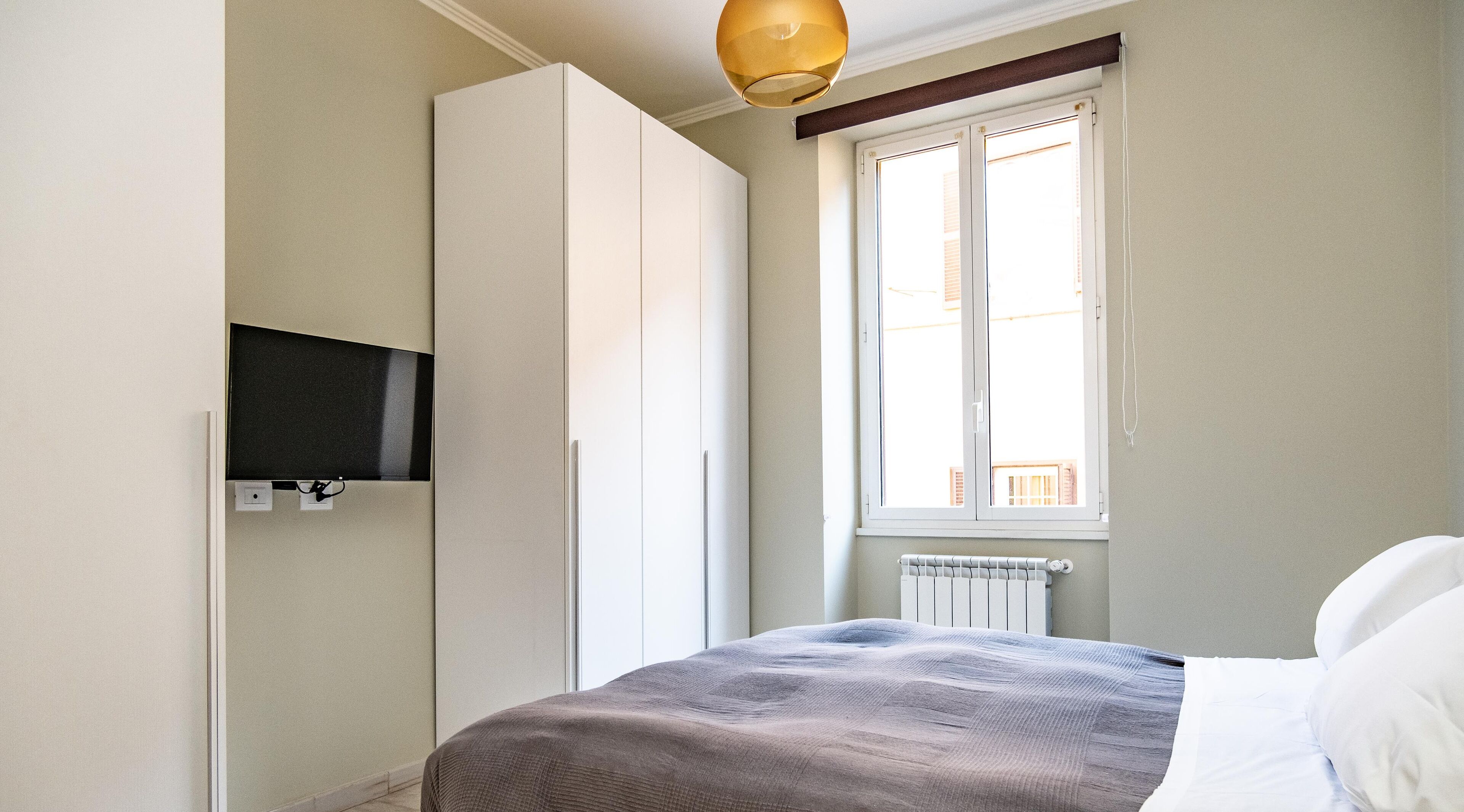 A bedroom with a bed, a wardrobe, a television, and a window with a view of buildings.