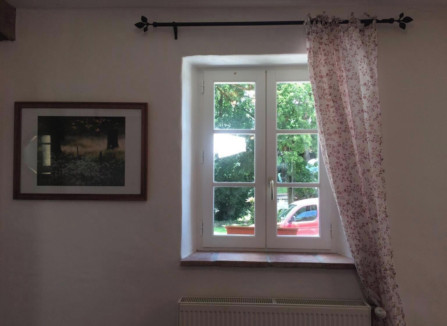 Een kamer met een raam naar het terras, een radiator en een lijst met een afbeelding aan de muur.