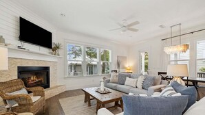 A living room with a fireplace, comfortable seating, a flat-screen TV, and large windows. - GORGEOUS Highland Parks w LSV & Bikes! Loaded 30A Home! (Santa Rosa Beach)