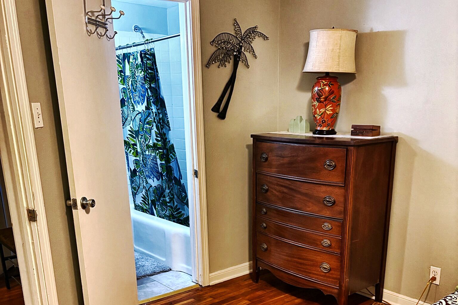 Une pièce comprenant une commode en bois, une lampe et une porte donnant sur une salle de bain avec un rideau de douche.