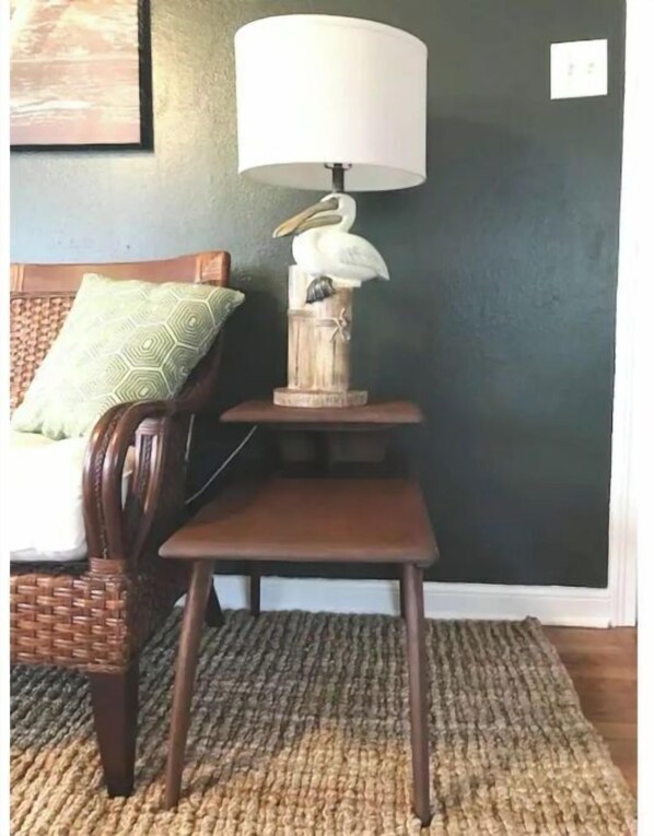 Una habitación con una silla de madera, una pequeña mesa de madera, una lámpara y un estante con un pájaro decorativo.