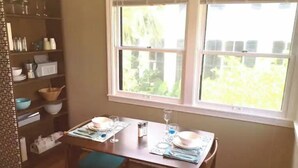 A dining area with a table set for two, a window with a view of greenery, and a bookshelf with various items. - Sealy Suites D  Historic East End - Walk to Beach, UTMB & Strand  (Galveston)