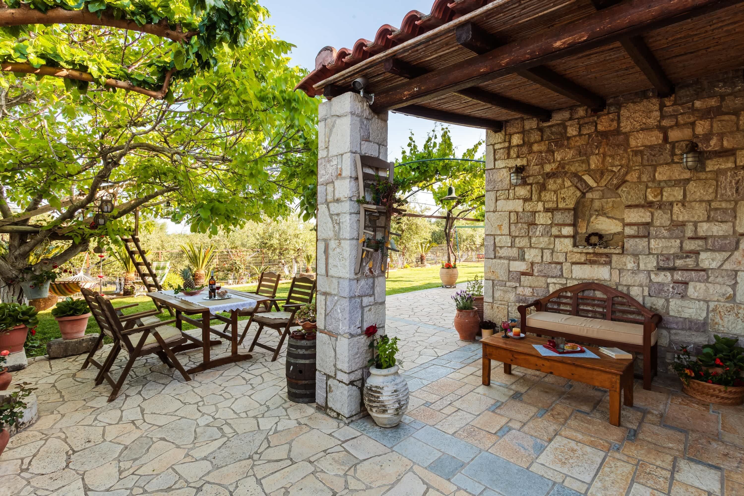 A stone patio with a wooden table and chairs, a bench, and potted plants.