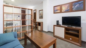 A living room with a wooden coffee table, a blue sofa, a TV on a wooden stand, and a glass partition with a kitchen area visible behind it. - LA CICER BEACH APARTMENT ll (Palmas De Gran Canaria, Las)