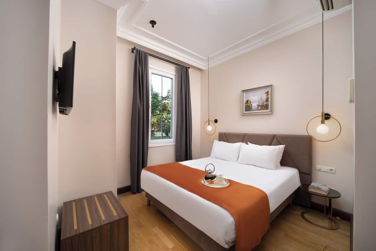 A hotel room with a large bed, a small side table, a wall-mounted TV, and a window with curtains.