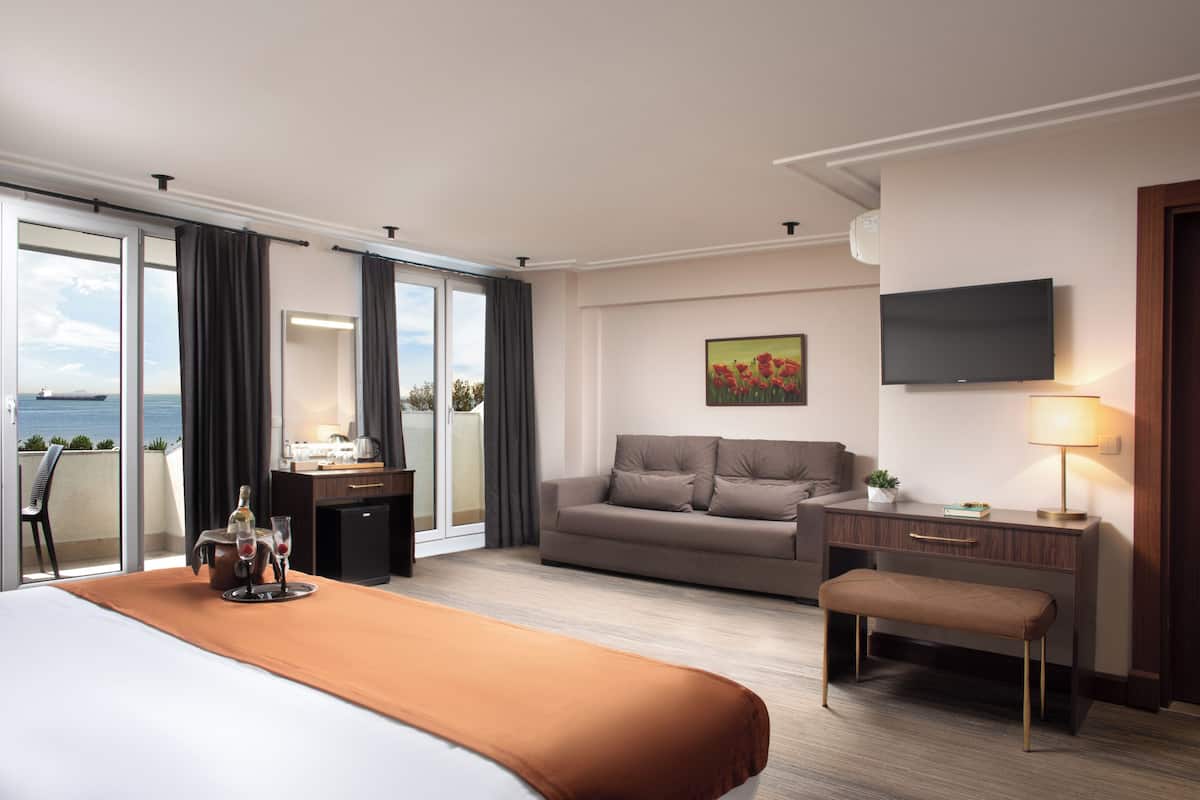 A hotel room with a bed, a sofa, a desk, a television, and a view of the sea.