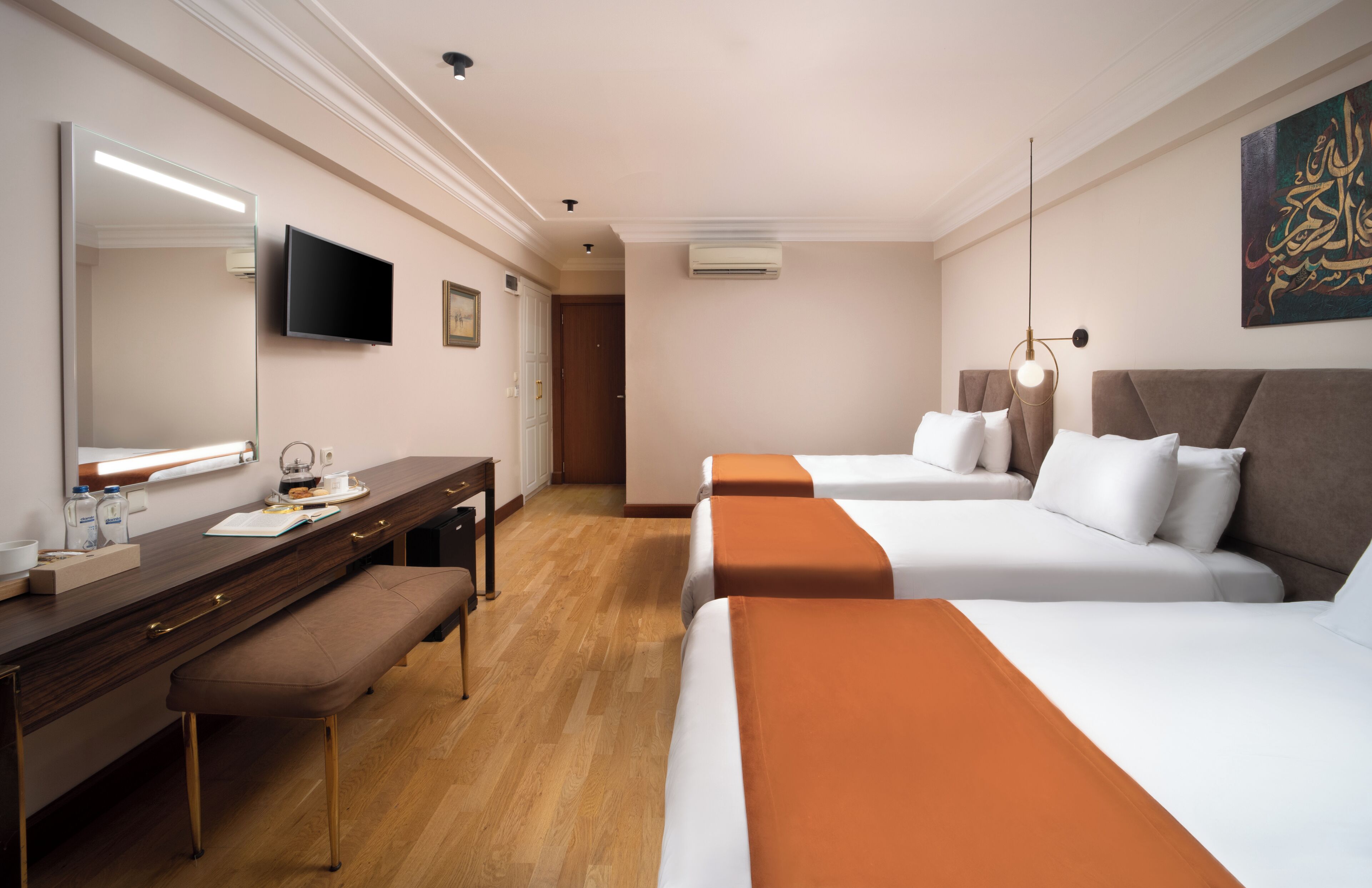 A hotel room with two beds, a desk with a mirror, a bench, a television, and a painting on the wall.