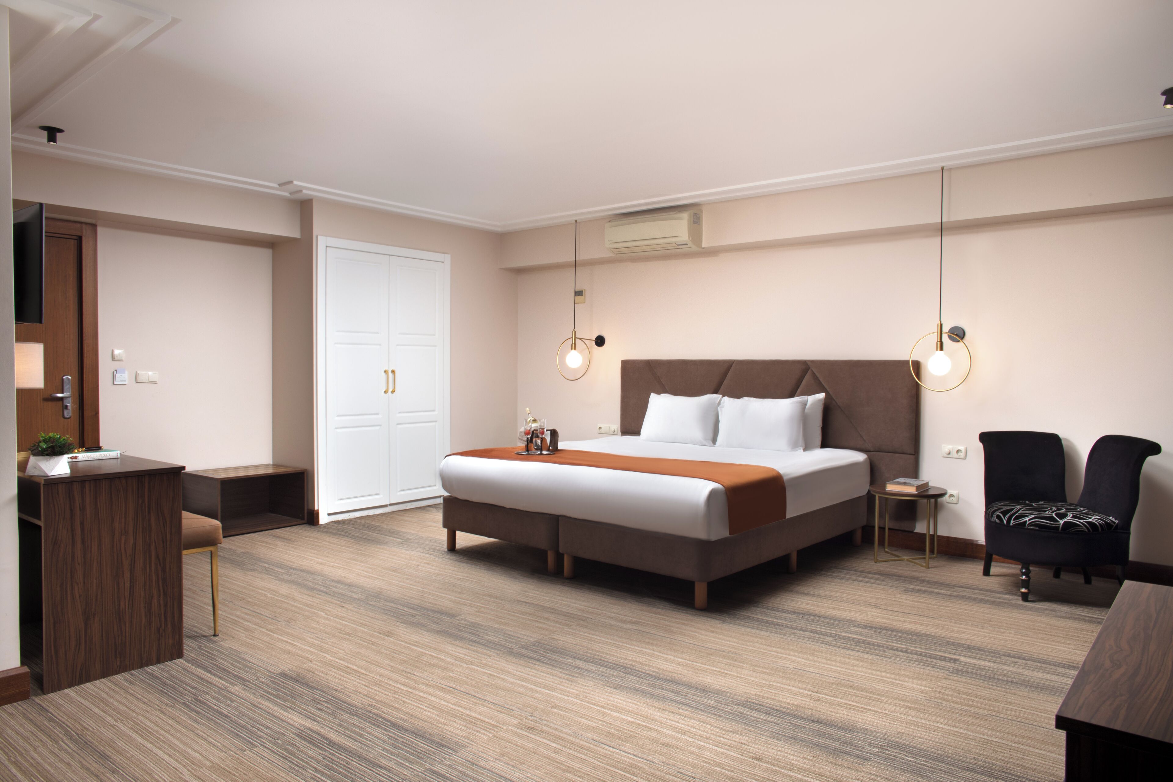 A modern hotel room with a large bed, a desk, and a chair. There are two pendant lights above the bed.
