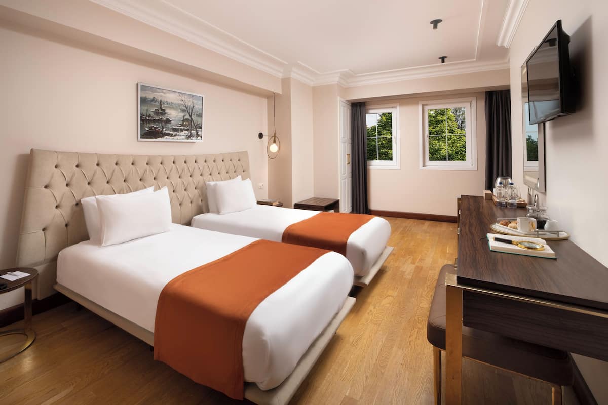 A hotel room with two beds, a wooden desk, a television, and a window with a view of greenery.
