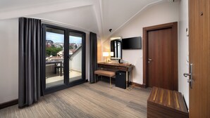 Habitación de hotel con un amplio ventanal corredero que da a un balcón, un escritorio de madera, un pequeño banco, un televisor de pantalla plana montado en la pared y un armario de madera.
