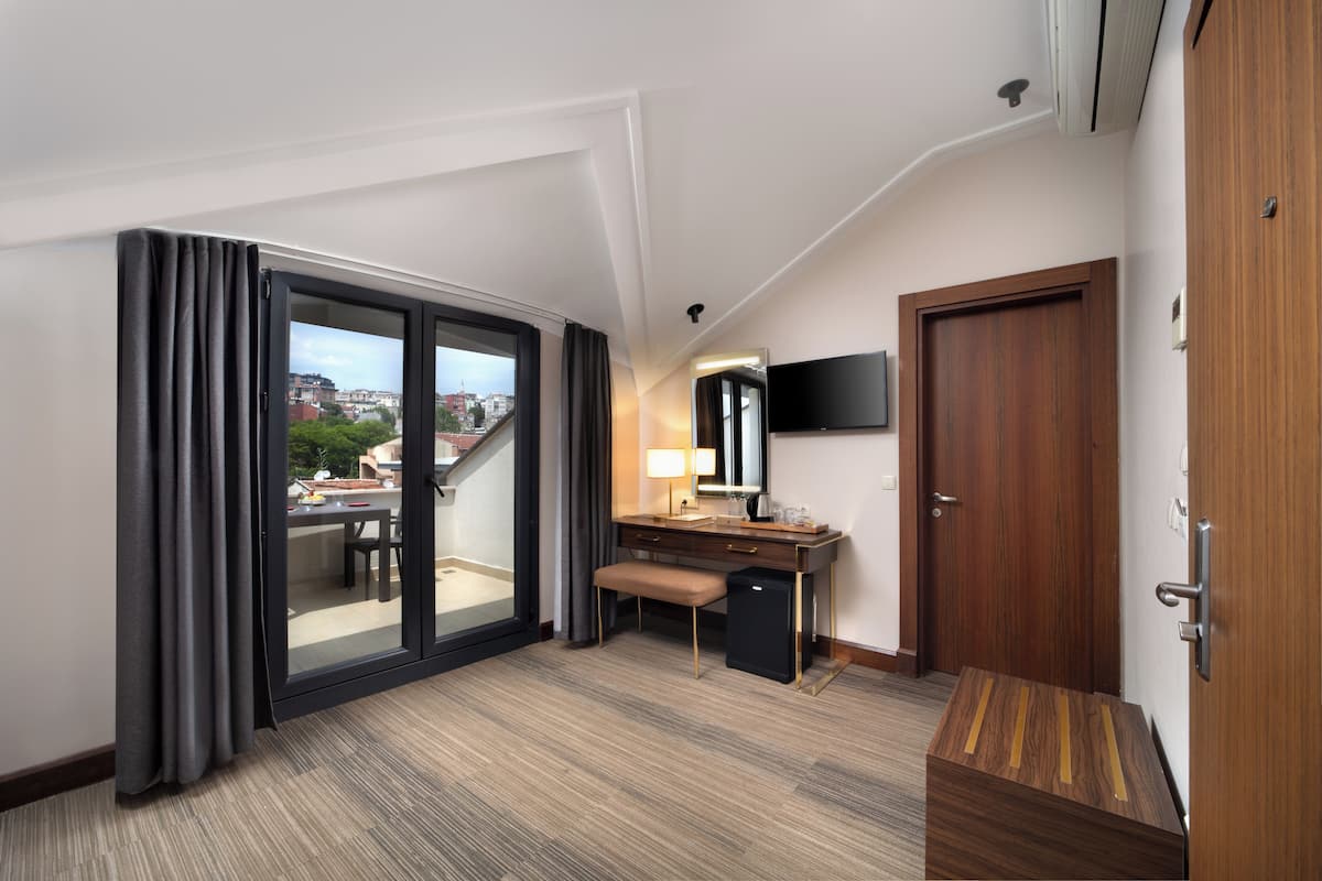 A hotel room with a large sliding glass door leading to a balcony, a wooden desk, a small bench, a flat-screen TV mounted on the wall, and a wooden wardrobe.