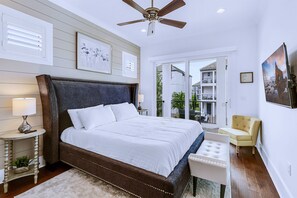 Une chambre avec un grand lit, un ventilateur de plafond, une table de chevet avec une lampe, une petite chaise et une vue sur le balcon extérieur.
