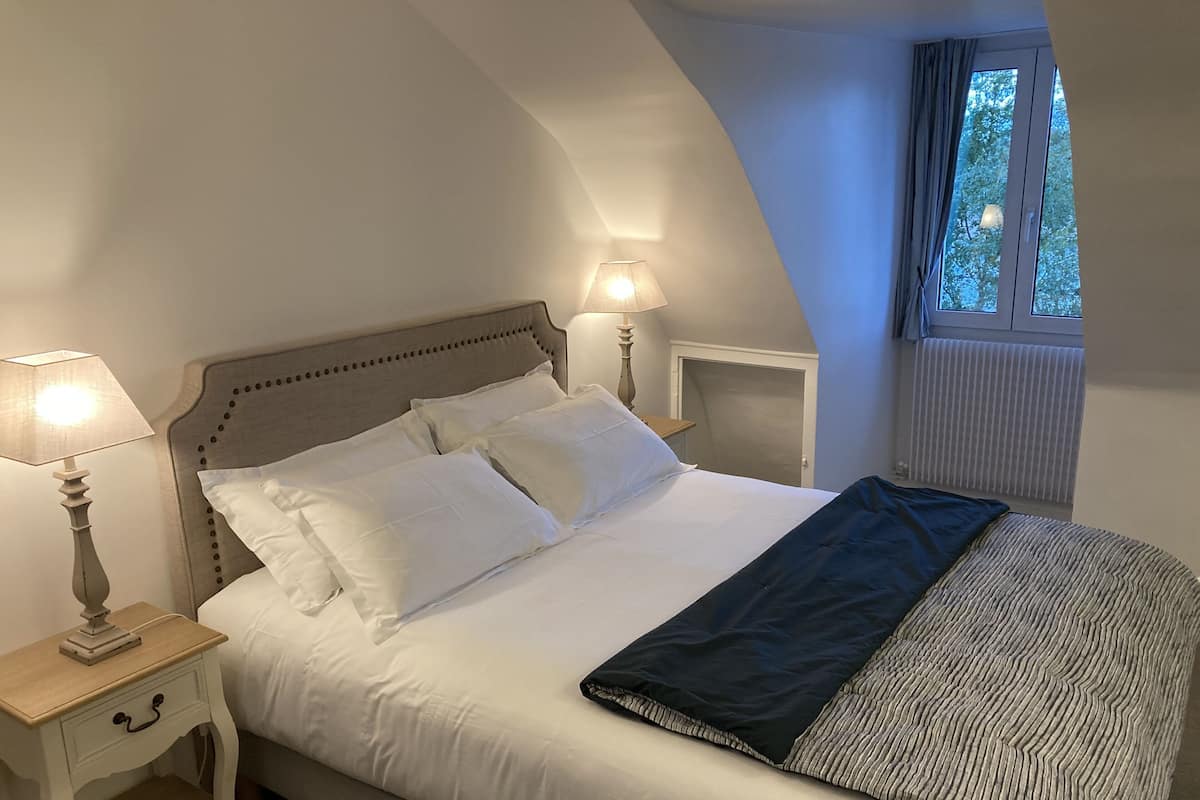 A neatly made bed with white linens and a dark blanket, a bedside table with a lamp, and a window with curtains.