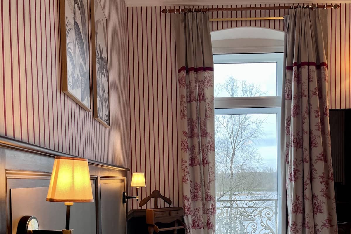 A hotel room with a bed, two pillows, a nightstand, a lamp, a chair, and a window with curtains.