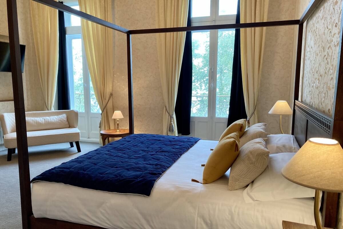 A four-poster bed with a blue bedspread, white linens, and beige pillows. There is a bedside table with a lamp, a chair, and a view of greenery through the window.