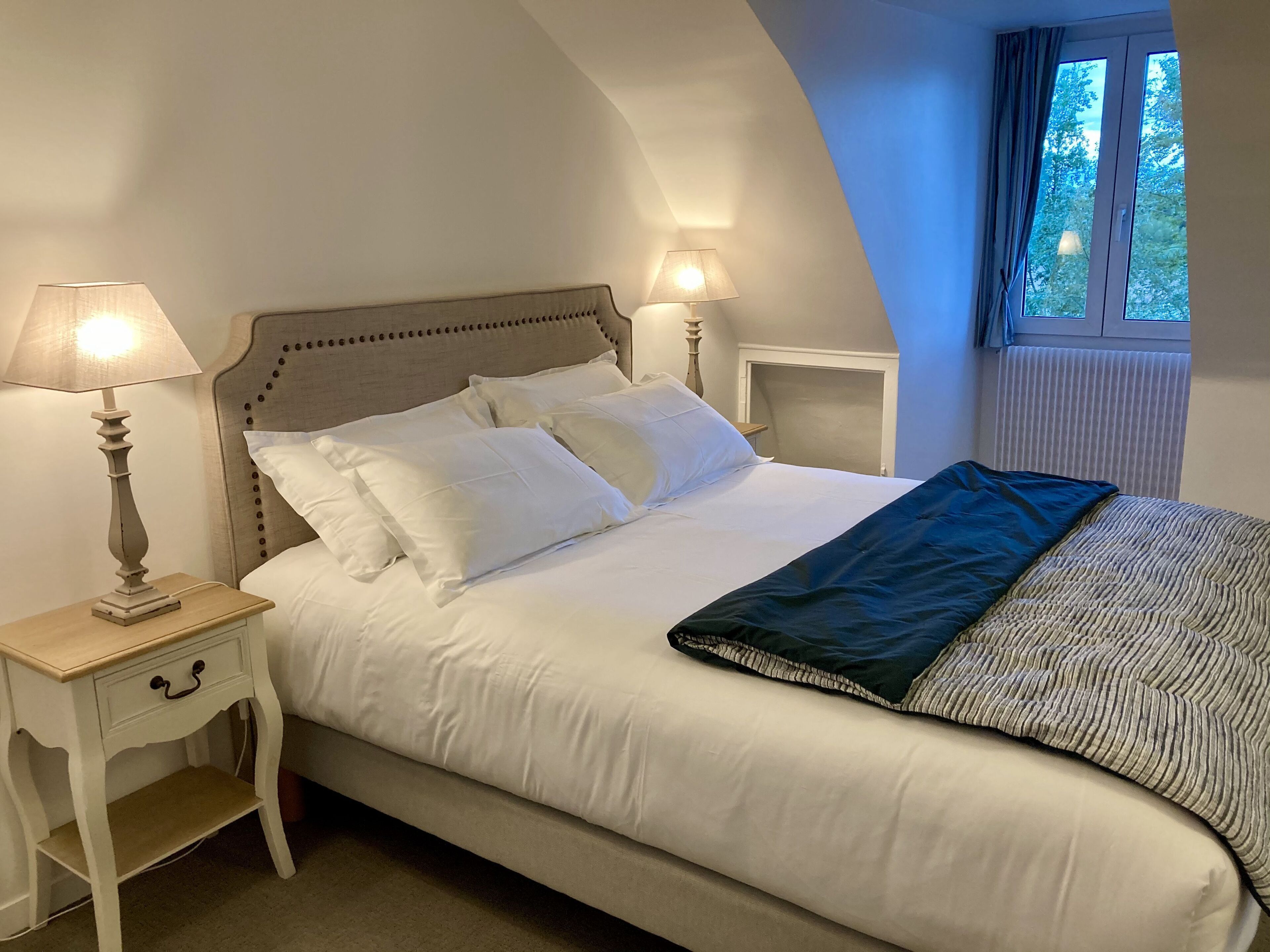 Een net opgemaakt bed met witte lakens en een donkere plaid, twee bedlampen en een raam met gordijnen.