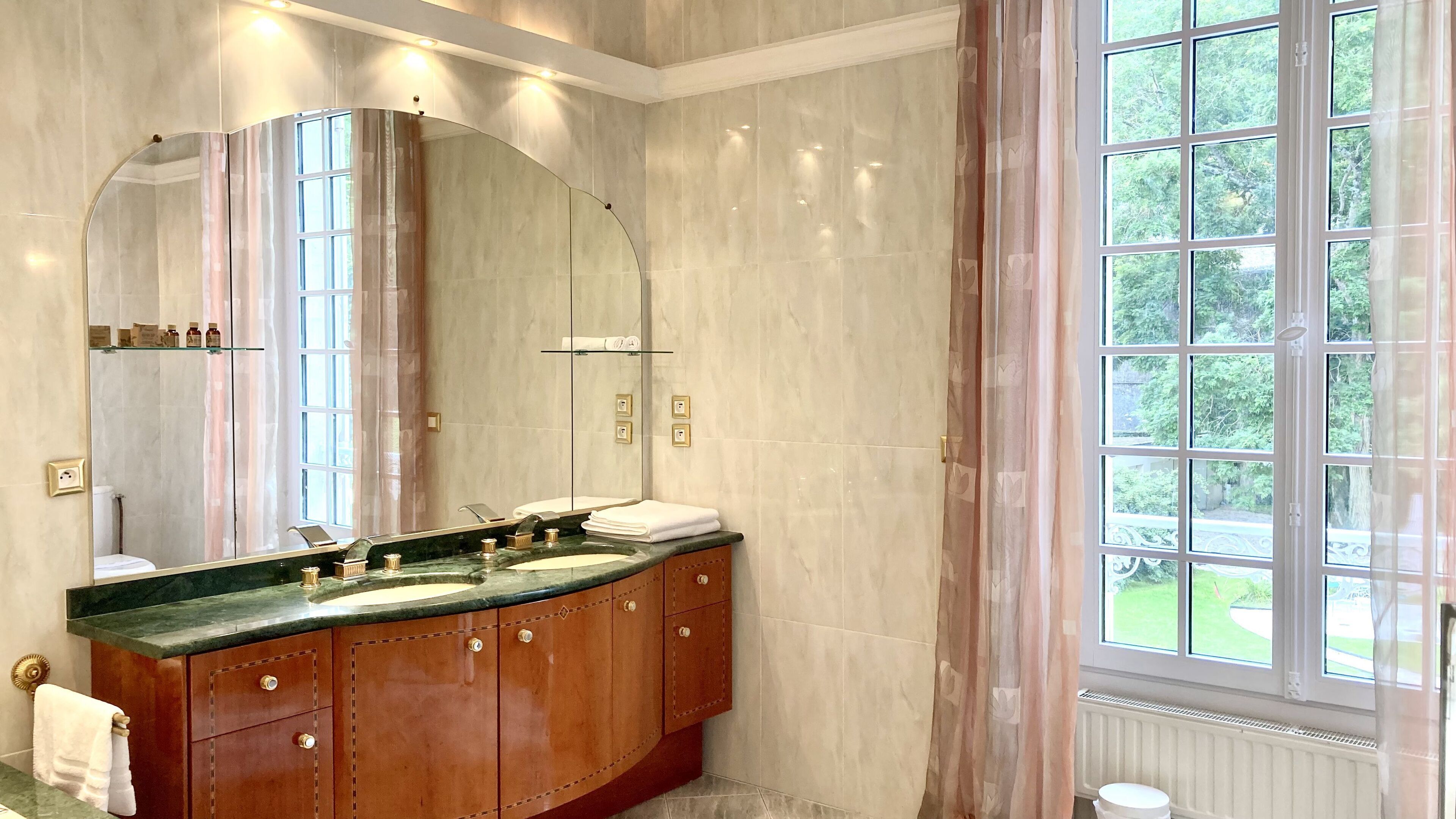 A bathroom with a large mirror, two sinks, wooden cabinets, and a window with a view of greenery.
