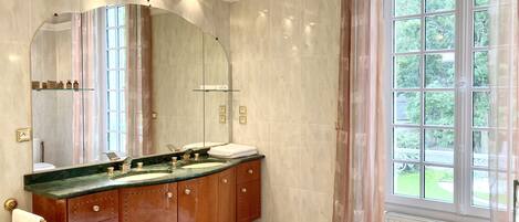A bathroom with a large mirror, two sinks, wooden cabinets, and a window with a view of greenery.