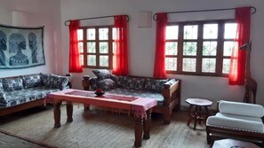 A living room with a sofa, a small table, and a chair. There are windows with red curtains and a painting on the wall. - Stay.Plus Malindi Apartment (Malindi)