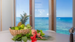 Une table sur laquelle se trouve un panier tressé rempli de fruits et de fleurs, un salier, et d’où l’on peut admirer l’océan à travers de grandes fenêtres.
