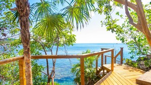 Une terrasse en bois dotée d’une balustrade, qui offre une vue imprenable sur une mer d’un bleu profond, le tout entouré d’une végétation luxuriante.