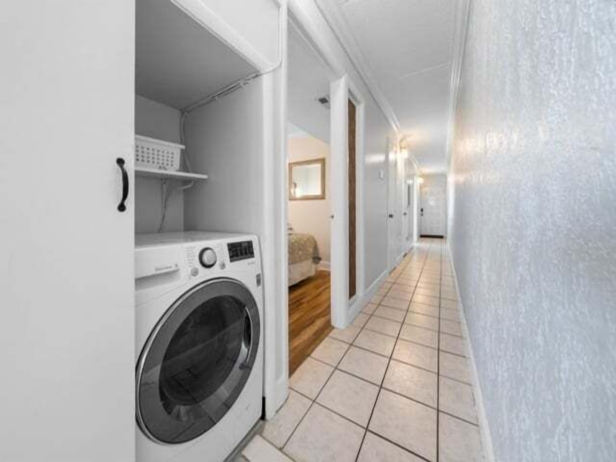 A laundry area with a washing machine, a shelf, and a bed in a hallway.