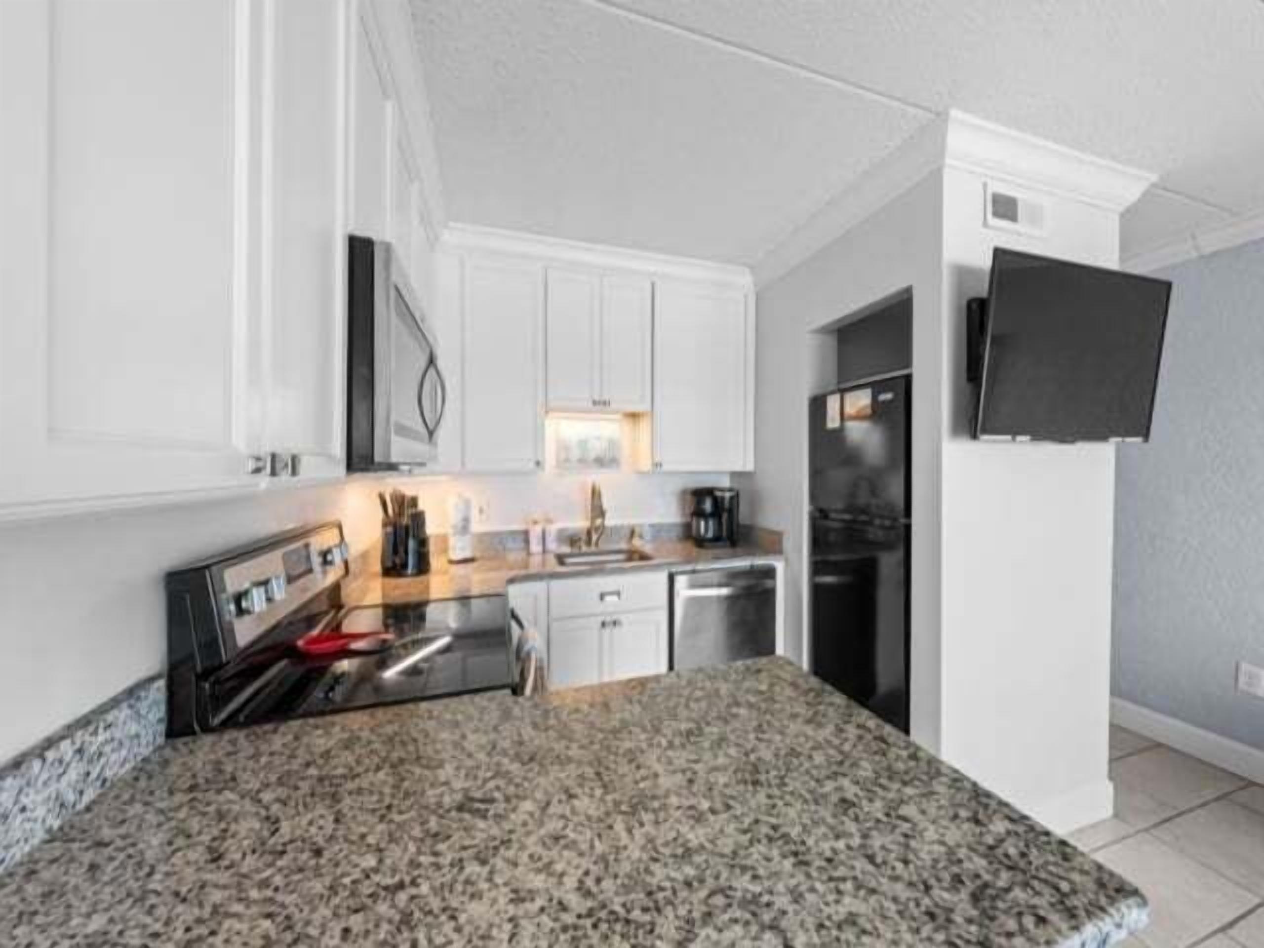 A modern kitchen with white cabinets, a granite countertop, a built-in oven, a coffee maker, and a flat-screen TV mounted on the wall.